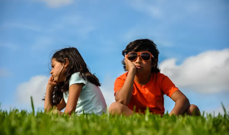 cuban indian brother sister boy girl sitting on grass angry bored hand to chin frustrated back to back blue sky white clouds orange shirt blue shirt hair blowing thinking
