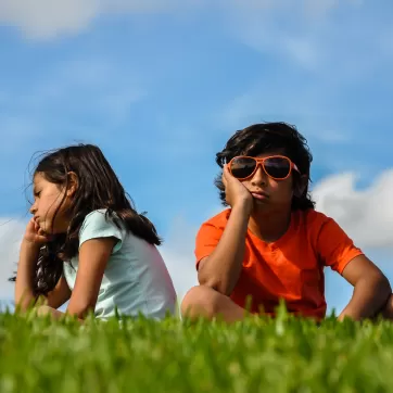 cuban indian brother sister boy girl sitting on grass angry bored hand to chin frustrated back to back blue sky white clouds orange shirt blue shirt hair blowing thinking