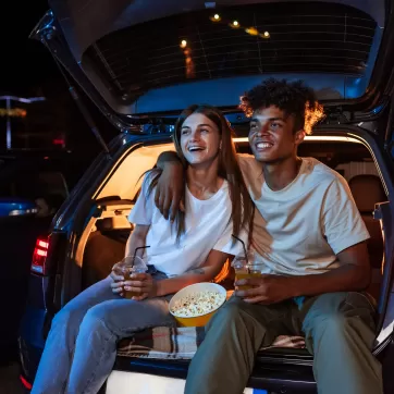 Diverse young couple having romantic date. Cheerful guy and his girlfriend watching a movie, sitting together in car trunk in front of a screen in an open air cinema