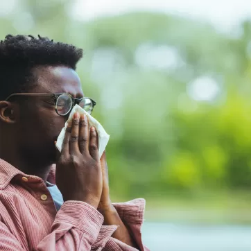 Allergic black man blowing on wipe in a park on spring season. Man with allergy or cold, blowing his nose with a tissue, looking miserable unwell very sick, isolated outside green trees background